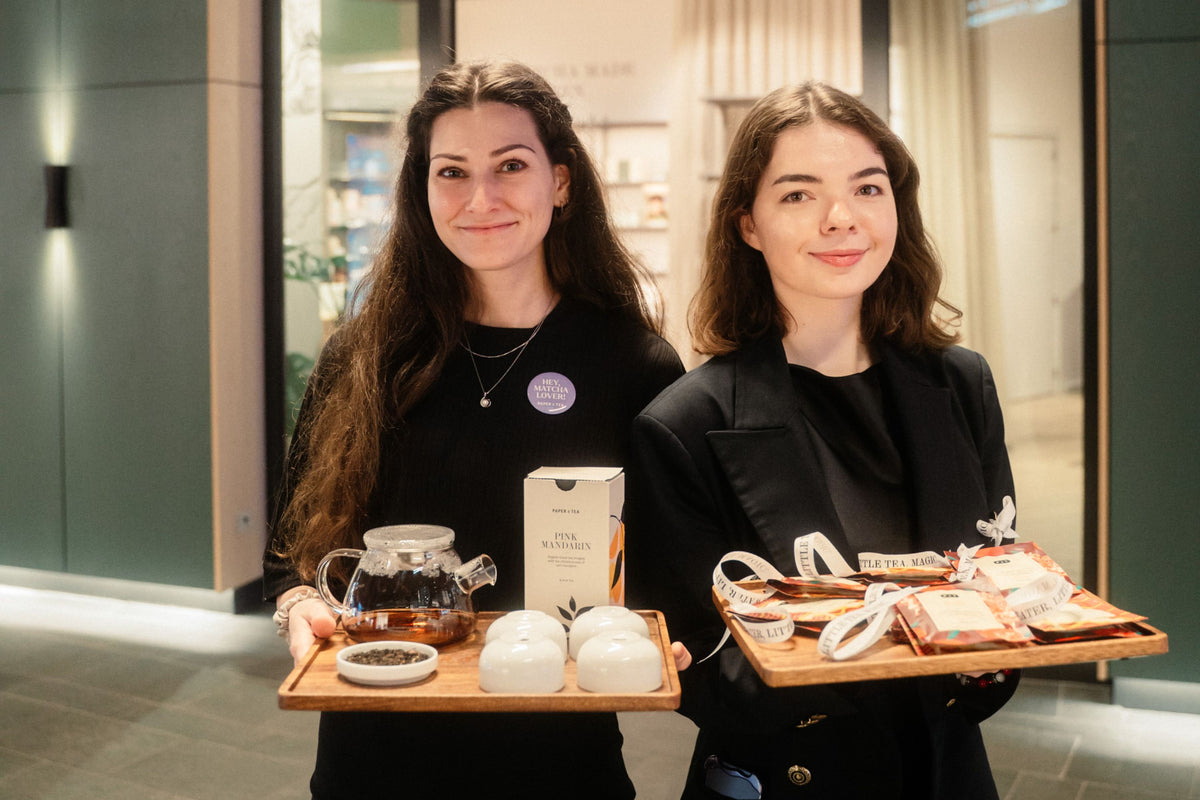 Zwei lächelnde Frauen präsentieren Teeprodukte von PAPER & TEA in einem modernen Teegeschäft.