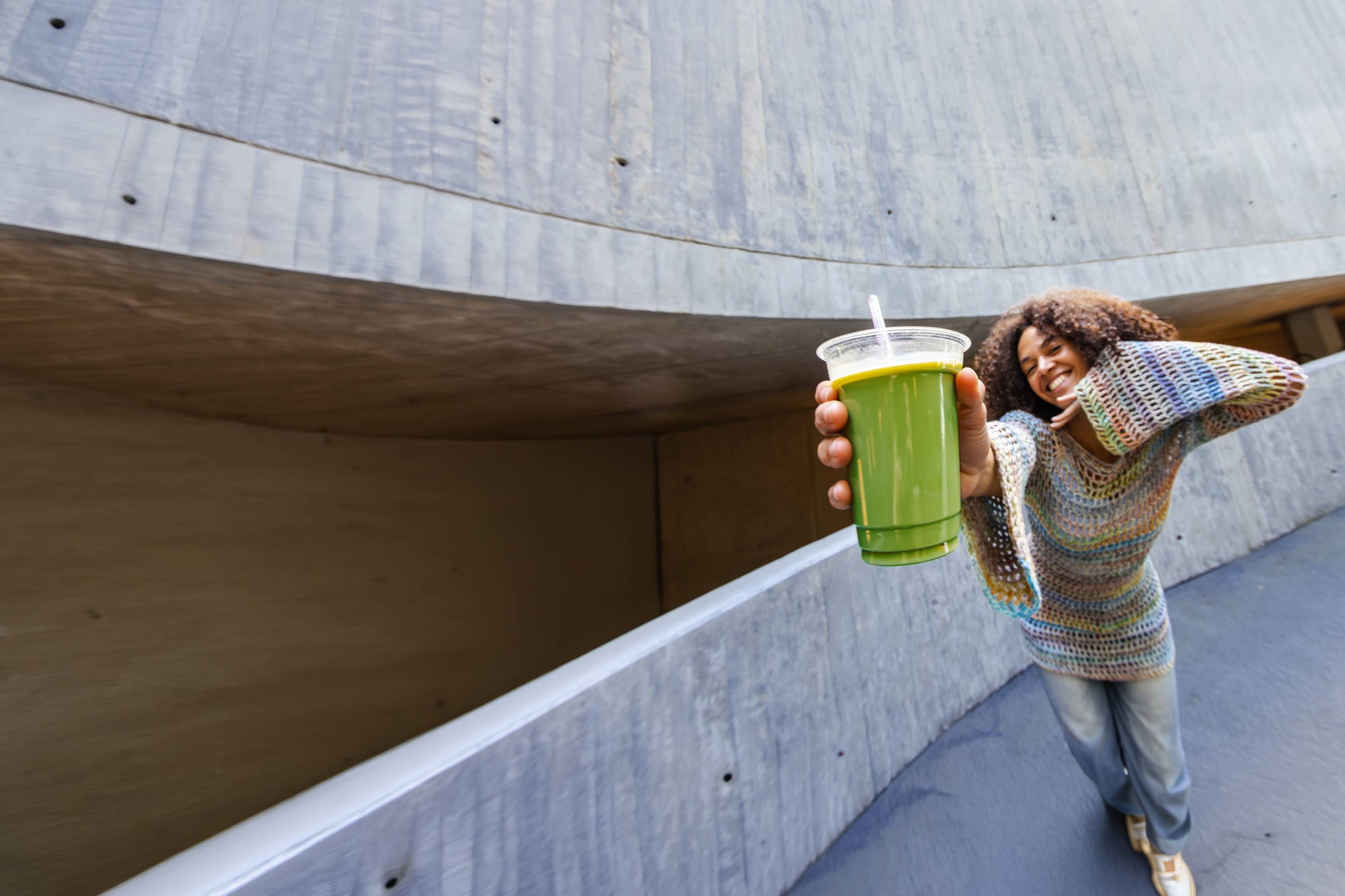 Junge Frau mit lockigem Haar im bunten Strickpullover und Jeans hält grünen Smoothie vor Betonwand.