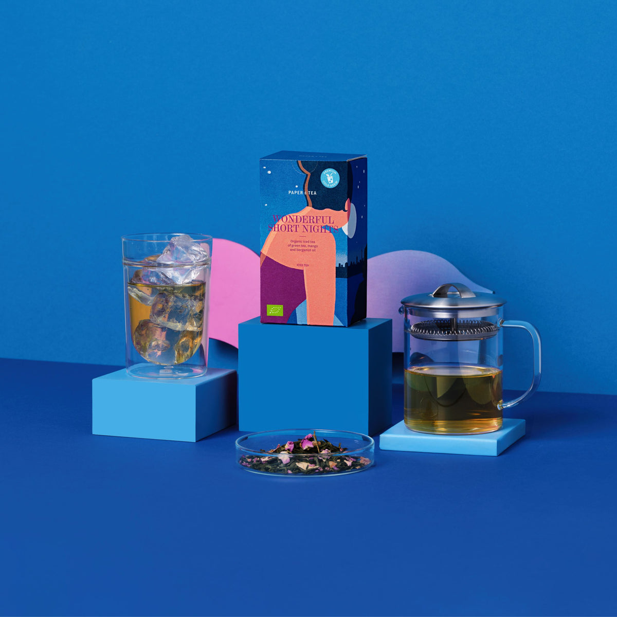 Bunte Verpackung von PAPER & TEA WONDERFUL SHORT NIGHTS Eistee neben Glas mit Eis, Teekanne und Teeblättern.
