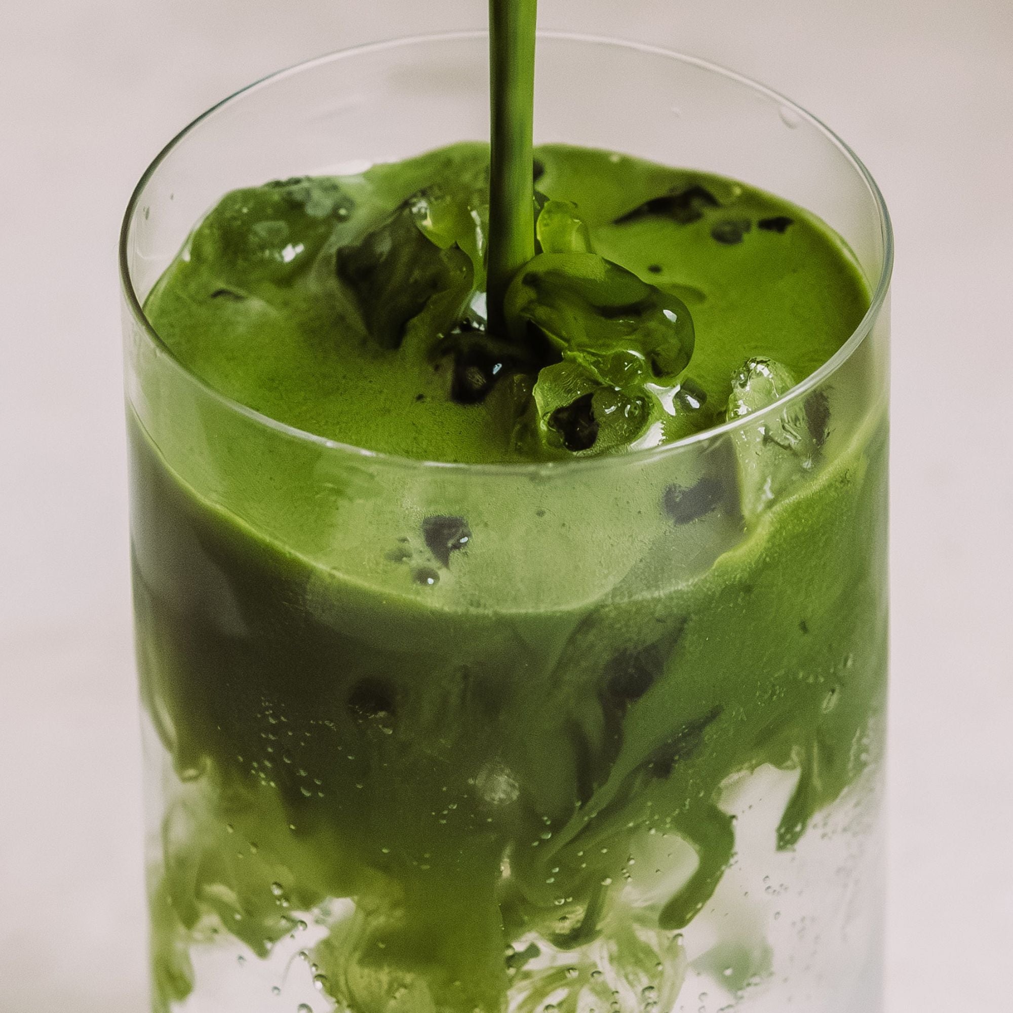 Klares Glas mit Eiswürfeln, in das leuchtend grüner Tee mit dynamischen Wirbeln gegossen wird.