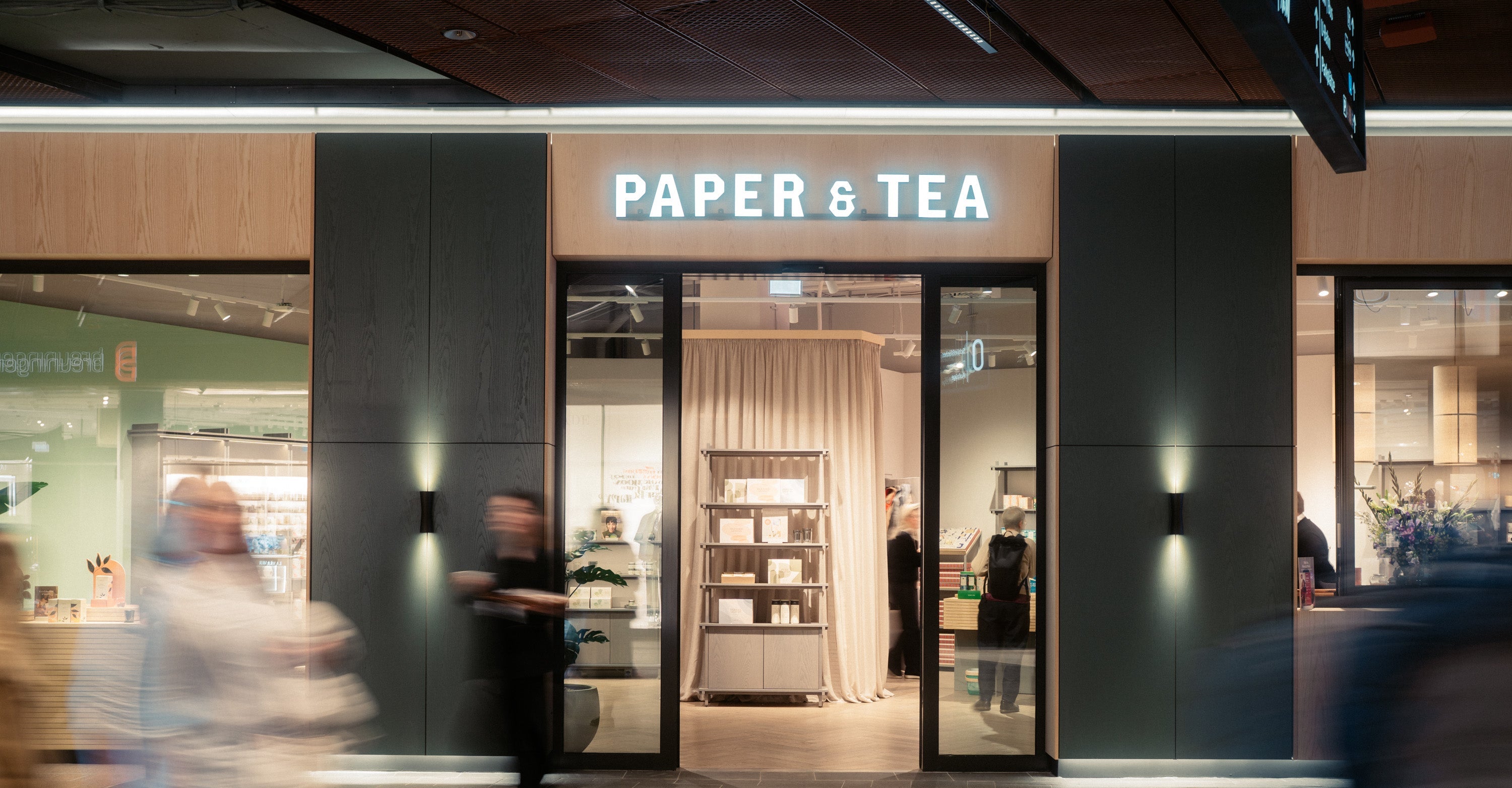 Eingang des modernen PAPER & TEA Ladens mit beleuchtetem Schild, Glasfront und warmem Licht.