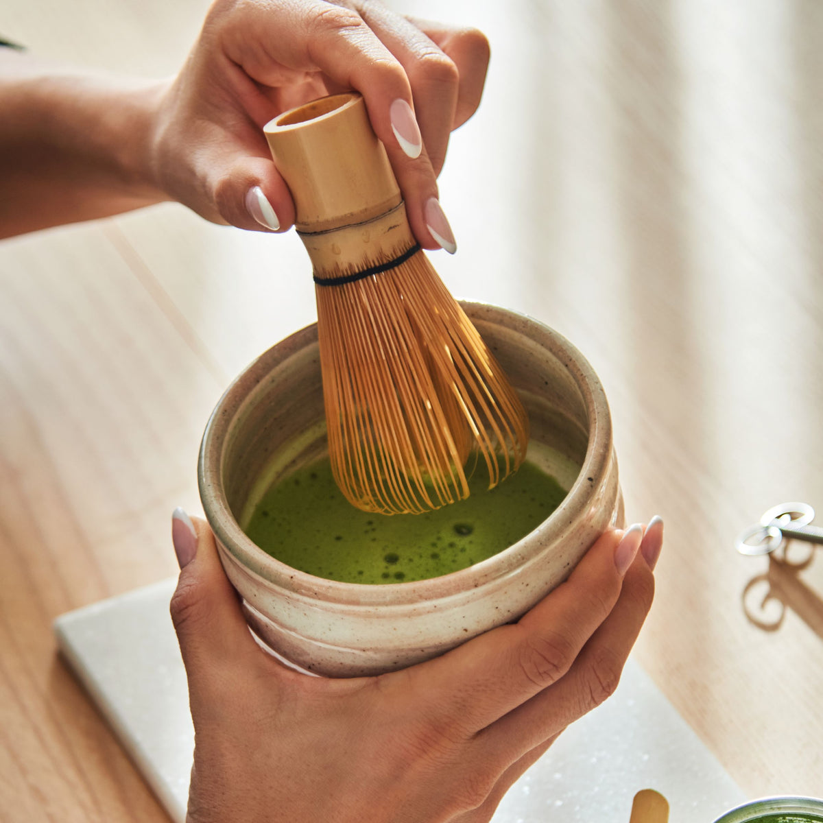 Hände bereiten Matcha-Tee mit Bambusbesen in rustikalem Keramikgefäß auf hellem Holztisch zu.