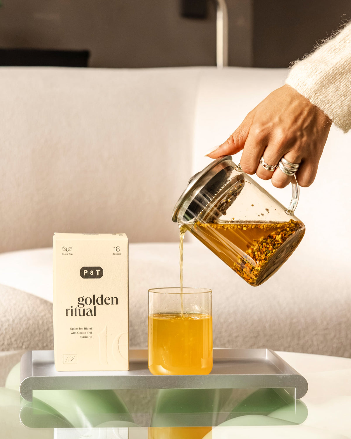 Beige P&T-Teebox Golden Ritual Gewürztee mit Kakaonibs und Kurkuma. Hand gießt Tee aus Glaskanne in Tasse.