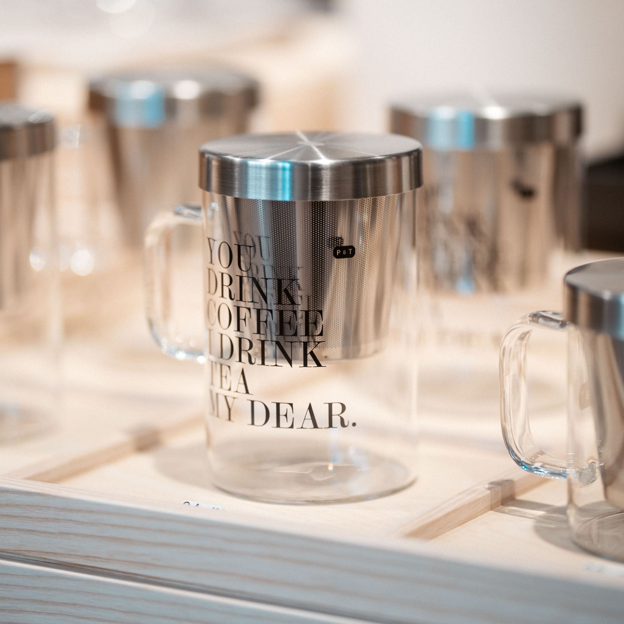 Glasbecher mit Edelstahldeckel und Sieb, bedruckt mit Kaffee- und Tee-Texten, auf Holztablett