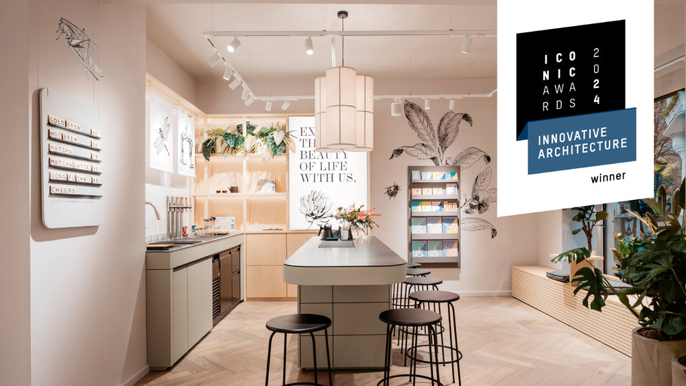 Moderner Teeladen mit zentraler Insel, schwarzen Stühlen, Teekannen, großer Pendelleuchte und botanischen Wandbildern.