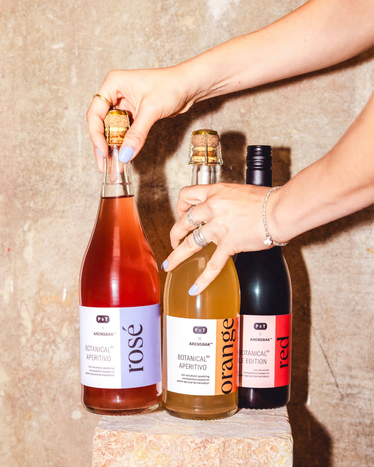 Zwei Hände mit lila Nagellack und Schmuck halten drei P&T Aperitivo-Flaschen in Lavendel, Orange und Rot auf einem Steinsockel.